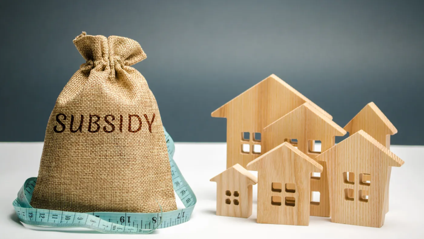 「SUBSIDY」と書かれた布袋と、木製の家の模型、メジャー