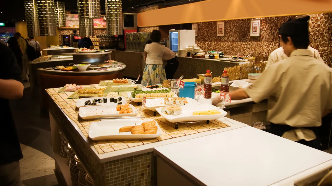寿司店のカウンター。白木の台に握りの皿が並び、奥で職人が寿司を握っている