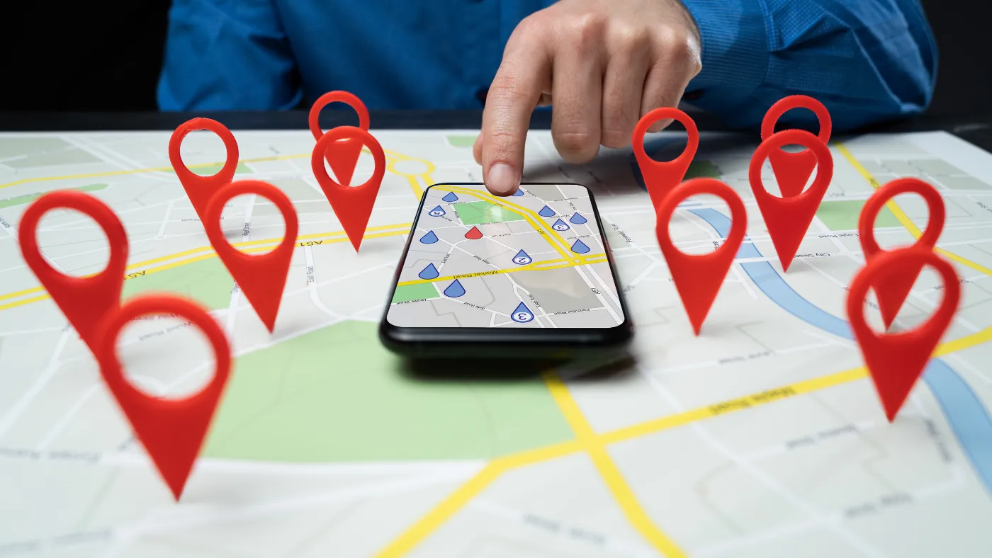 机に広げた地図の上に赤い位置ピンがいくつも立ち、中央に置かれたスマートフォンの地図を指で押している手元