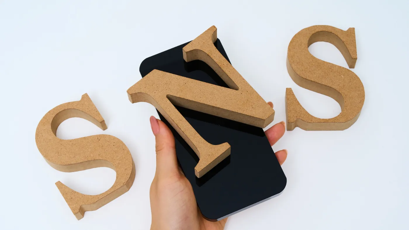 木でできた「S」「N」「S」の文字と、その「N」を載せたスマートフォンを持つ手