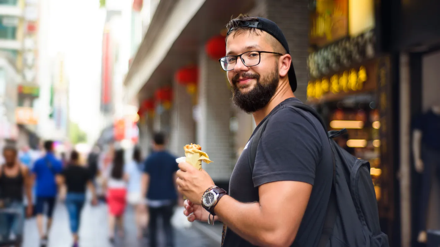 赤いちょうちんが下がる商店街で、リュックを背負った男性が食べ歩きのクレープを手に振り返っている