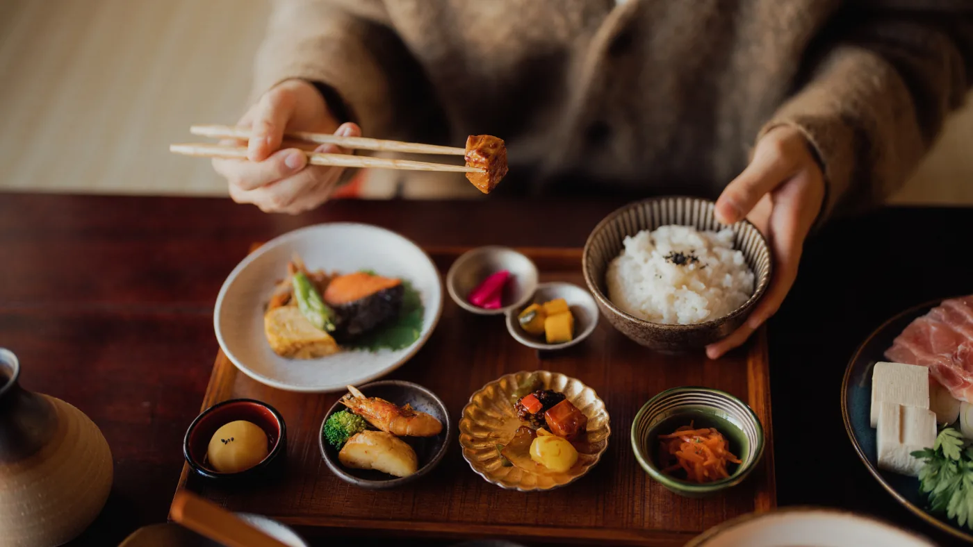 お盆に小鉢がいくつも並んだ和定食を箸で食べている手元