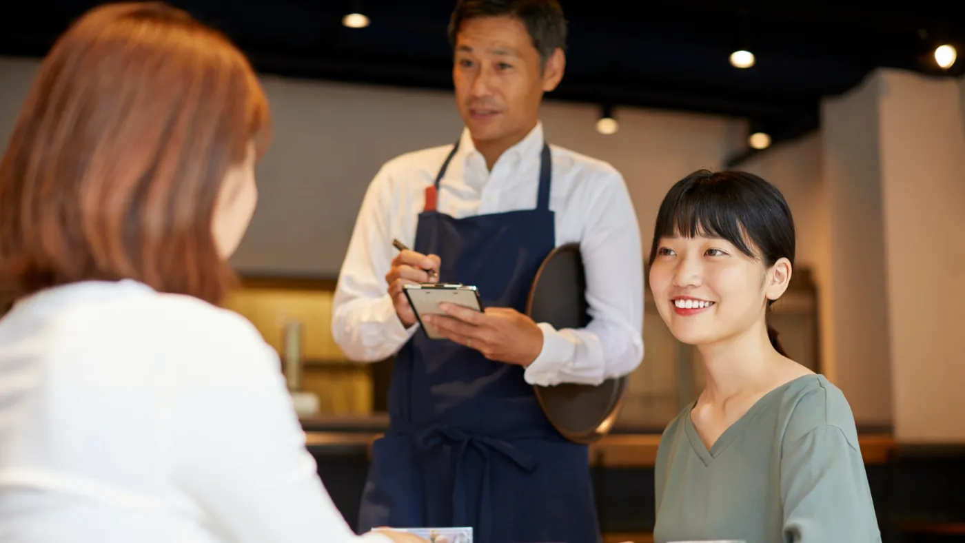エプロン姿の男性店員が伝票を手に、席の女性客2人から笑顔で注文を聞いている